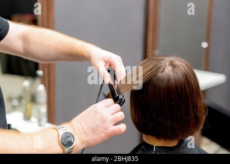Friseurhände sind Glätten Haar der Frau mit Haarglätter Werkzeug im Friseursalon, Rückansicht. Stockfoto
