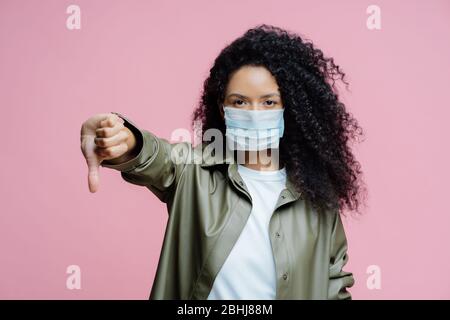 Bild von ernsten lockig behaarte junge Frau trägt medizinische Maske, hält den Daumen nach unten, zeigt Abneigung Geste, vermeidet Verbreitung von Corona-Virus, kümmert sich um h Stockfoto