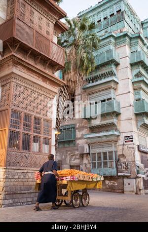 Jeddah / Saudi Arabien - 16. Januar 2020: Mann schiebt einen mit Orangen beladenen Wagen mit historischen bunten Gebäuden im Hintergrund in Al-Balad Stockfoto