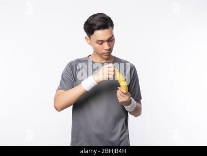 Junger asiatischer Sportmann in grauem T-Shirt und weißen Armbändern, die eine Banane auf weißem Hintergrund isoliert abblätternd. Stockfoto