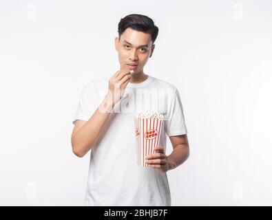 Junger asiatischer Mann in weißem T-Shirt hält und essen Popcorn mit glücklichem Gesicht isoliert auf weißem Hintergrund. Stockfoto