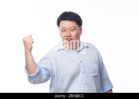 Aufgeregt asiatischen Fat man in blauem Hemd heben seine Faust mit glücklich erfreut Gesicht, feiern Erfolg isoliert auf weißem Hintergrund. Stockfoto