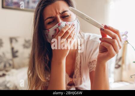 Schwanger während der Coronavirus Covid-19 Pandemie. Kranke Frau in Maske Überprüfung positive Schwangerschaftstest zu Hause husten. Stockfoto