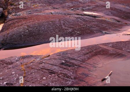 Nahaufnahme des glazial polierten Felsens, beleuchtet vom Winteruntergang und orangefarbenen Wolken, die sich im stehenden Wasser am Trondheimsfjord, Norwegen, spiegeln Stockfoto