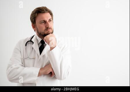 Porträt des männlichen Arztes mit Stethoskop in medizinischer Uniform, die Hand unter dem Kinn haltend, zweifelhaft auf einem weißen isolierten Hintergrund posiert Stockfoto