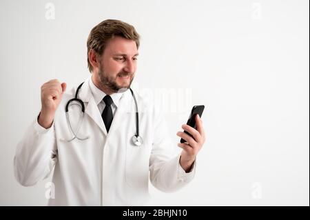Porträt des männlichen Arztes mit Stethoskop in medizinischer Uniform mit blonden Haaren, Smartphone halten, SMS Nachrichten glückliches Gesicht posiert auf einem weißen Isolat