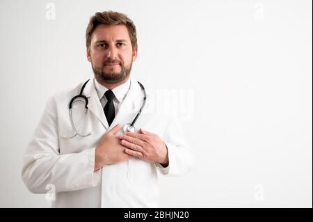 Portrait des männlichen Arztes mit Stethoskop in medizinischer Uniform hält ihre Hände auf der Brust nahe ihrem Herzen posiert auf einem weißen isolierten Hintergrund Stockfoto