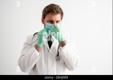 Portrait des männlichen Arztes mit Stethoskop in medizinischer Uniform arrangiert seine Schutzmaske posiert auf einem weißen isolierten Hintergrund Stockfoto