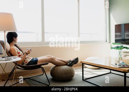 Junge Frau sitzt vor dem Fenster in ihrer Wohnung Stockfoto