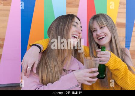 Zwei fröhliche junge kaukasische Frauen stoßen mit einem Drink an die bunte Wand einer Stadtbar Stockfoto