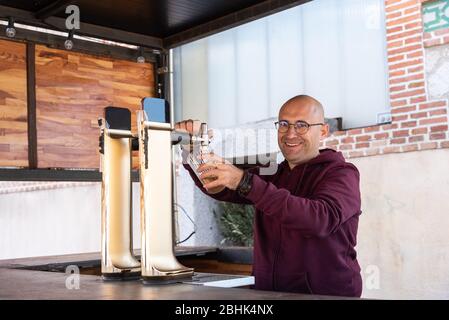 Kaukasischer Erwachsener Mann ohne Haare und trägt Gläser funktioniert lächelnd und glücklich serviert ein Bier in einer Bar. Stockfoto