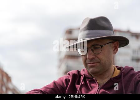 Kaukasischer Mann mit Brille und Hut depressiv und traurig in einem einsamen Rollstuhl in einem Park mit Himmel und einigen Gebäuden im Hintergrund mit Raum Stockfoto