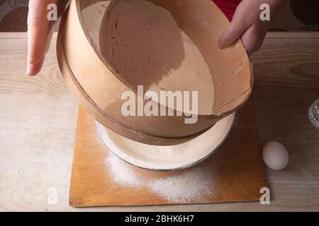 Große Schüssel, Schneidebrett, Sieb zum Sieben von Mehl auf dem Tisch. Weibliche Hände sieben Mehl in der Küche Stockfoto