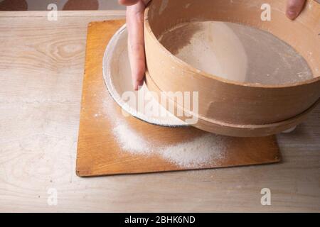 Große Schüssel, Schneidebrett, Sieb zum Sieben von Mehl auf dem Tisch. Weibliche Hände sieben Mehl in der Küche Stockfoto