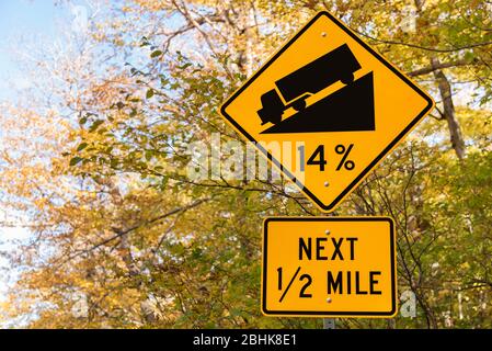 Steiles Schild mit Autunalbäumen im Hintergrund. Fahrer werden vor einer steilen 14% Steigung auf einer Bergstraße gewarnt.
