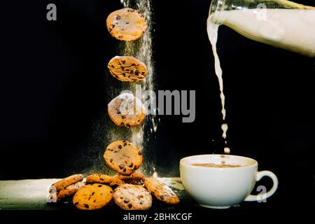 Tee Und Kekse. Kekse oder Kekse fallen auf einen Haufen von Cookies neben einer Tasse Tee auf schwarzem Hintergrund. Milch wird in eine Tasse Tee gegossen. Stockfoto