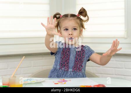 Ein kleines Mädchen in einem blauen Kleid malt am Fenster und zeigt, was für saubere Hände sie noch hat. Bleiben Sie zu Hause, Virus Covid-19, Selbstisolation, Isolation Stockfoto