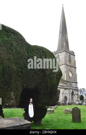 Blass junge Dame in flauschigen Wintermantel und Schal auf dem Friedhof einer traditionellen englischen Kirche stehen: eibenhecke, Kirchturm und kaltes graues Licht Stockfoto