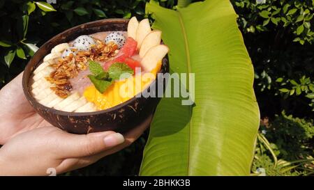 Die Hände der Frau halten bunte Smoothie Schüssel mit tropischen Früchten mit Granola auf Grün Dschungel Hintergrund. Vegetarisches Frühstückskonzept Stockfoto