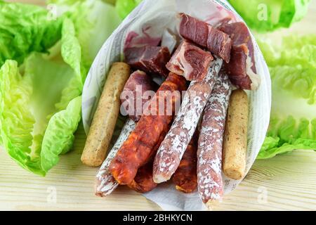Sortiment von Fuet und Chorizo Würstchen, Scheiben von Jamon und Brotsticks in Öko Papiertüte. Grüner frischer Salat auf dem Holztisch. Mittelmeer fas Stockfoto