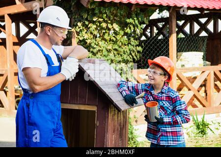 Vater und Sohn arbeiten zusammen in einer Holzwerkstatt. Sie konzentrieren sich auf den Bau eines hölzernen Hundehauses, der Sohn sitzt darin mit einer Farbe Stockfoto