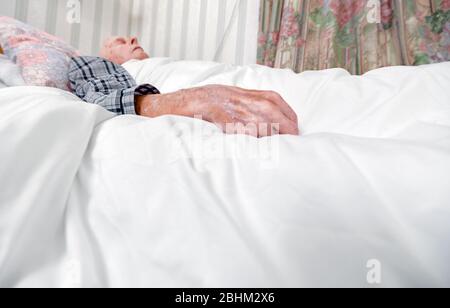 Nahaufnahme seiner Hand im Vordergrund, mit Pyjama in einem Krankenhausbett unten in seinem Haus. Stockfoto