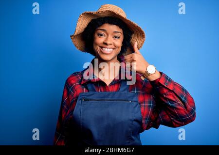 Junge afro amerikanische Afro Farmer Frau mit lockigen Haaren tragen Schürze und Hut tun glücklich Daumen hoch Geste mit der Hand. Genehmigen eines Ausdrucks, der nach einem sucht Stockfoto