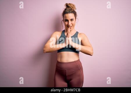 Junge schöne blonde Sportlerin Sport Sport tragen Sportbekleidung über rosa Hintergrund beten mit Händen zusammen bitten um Vergebung lächelnd conf Stockfoto