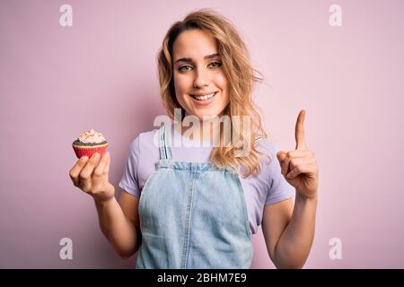 Junge schöne blonde Frau eatimg Schokolade Cupcake über isoliert rosa Hintergrund überrascht mit einer Idee oder Frage Zeigen Finger mit glücklichem Gesicht Stockfoto