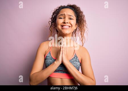 Schöne Sportlerin mit lockigen Haaren tun Sport tragen Sportbekleidung über rosa Hintergrund beten mit Händen zusammen bitten um Vergebung lächelnd c Stockfoto