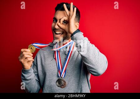 Junge gut aussehende erfolgreiche Mann Medaillen im Stehen über isoliert roten Hintergrund mit glücklichen Gesicht lächelnd tun ok Zeichen mit Hand auf Auge suchen throu Stockfoto