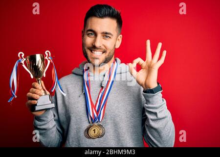 Junge gut aussehende erfolgreiche Mann mit Trophäe trägt Medaillen über rotem Hintergrund tun ok Zeichen mit Fingern, ausgezeichnetes Symbol Stockfoto