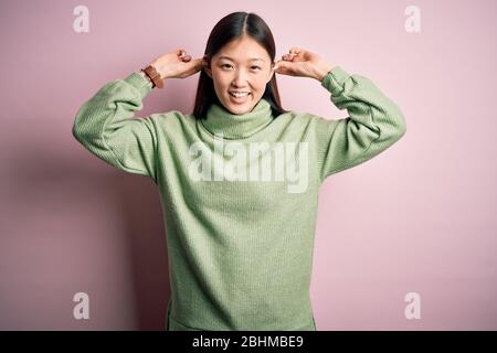 Junge schöne asiatische Frau trägt grünen Winter Pullover über rosa solierten Hintergrund lächelnd ziehen Ohren mit Fingern, lustige Geste. Probespiel Stockfoto