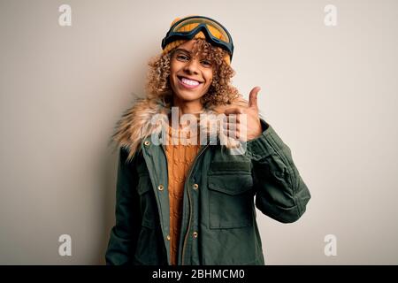 Junge afroamerikanische Skifahrer Frau mit lockigen Haaren tragen Schnee Sportbekleidung und Skibrille tun glücklich Daumen hoch Geste mit der Hand. Genehmigung von Ausdruckstexts Stockfoto