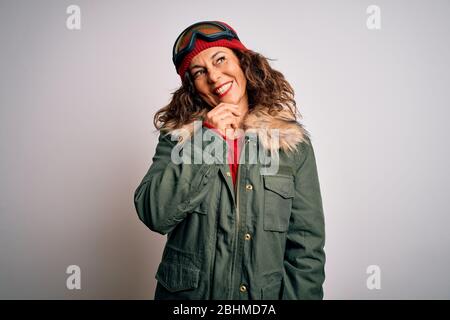 Mittlere Alter Skifahrer Frau trägt Schnee Sportbekleidung und Skibrille auf weißem Hintergrund schaut zuversichtlich auf die Kamera mit Lächeln mit verschränkten Armen und Stockfoto