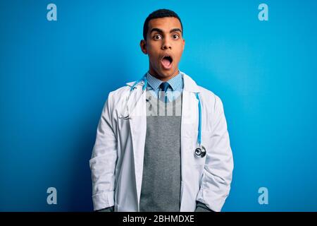 Schöner afroamerikanischer Arzt Mann trägt Mantel und Stethoskop über blauem Hintergrund im Schockgesicht, Blick skeptisch und sarkastisch, überrascht mit Stockfoto