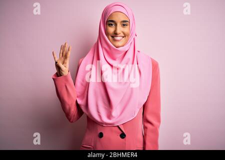 Junge schöne Brünette Geschäftsfrau trägt rosa muslimischen Hijab und Business-Jacke zeigt und zeigt mit den Fingern Nummer vier beim Lächeln Co Stockfoto