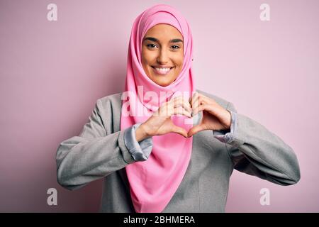 Junge schöne Brünette Geschäftsfrau trägt rosa muslimischen Hijab und Business-Jacke lächelnd in der Liebe zeigt Herz-Symbol und Form mit den Händen. Romant Stockfoto