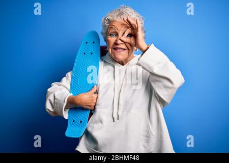 Senior schöne sportliche Frau hält Skate stehen über isoliert blauen Hintergrund mit glücklichen Gesicht lächelnd tun ok Zeichen mit Hand auf Auge suchen thro Stockfoto