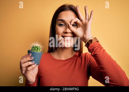 Junge schöne Brünette Frau, die kleine Kaktus Topf über gelben Hintergrund mit glücklichem Gesicht lächelnd tun ok Zeichen mit Hand auf Auge durch zu suchen Stockfoto