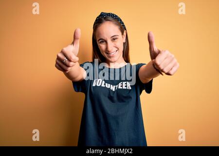 Junge schöne Frau trägt Freiwilligen T-Shirt tun Freiwilligenarbeit über gelben Hintergrund Genehmigung tun positive Geste mit der Hand, Daumen hoch Smilie Stockfoto