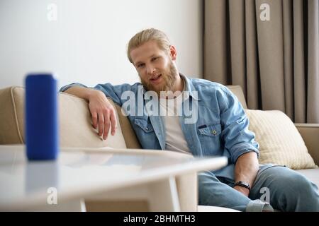 Junger Mann spricht, aktiviert tragbaren kabellosen Lautsprecher auf dem Tisch in Smart Home. Stockfoto