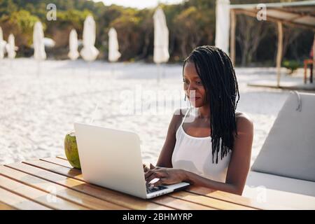 Hübsche junge Frau, die am Holztisch am Strand sitzt und am Laptop arbeitet Stockfoto