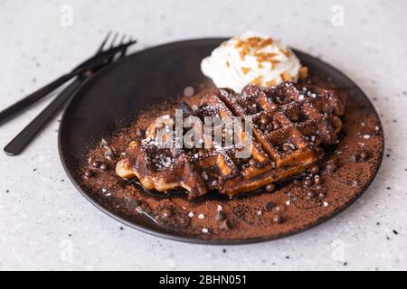 Leckere hausgemachte belgische Waffeltafel mit dunklem Schokoladensirup, Kakaopulver und Schokoladenchips, serviert mit Schlagsahne in brauner Farbe Platte. Stockfoto