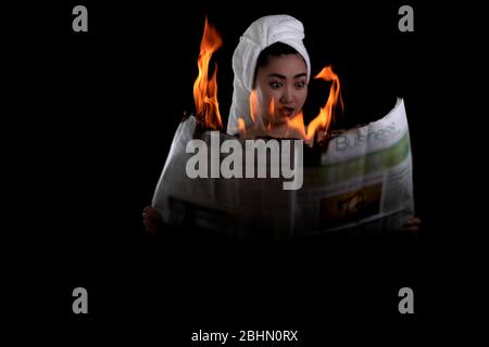 Junge schöne Asienfrau hält beleuchtet und liest eine brennende Zeitung auf dem schwarzen Hintergrund, Hot breaking News Konzept Stockfoto