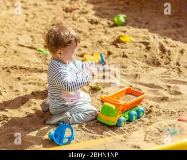 Kleines Baby Mädchen Kaukasischen Jungen spielt in der Sandbox mit einem Spielzeugauto. Ein Kind im gestreiften T-Shirt gießt Sand in einen orangefarbenen LKW mit Plastikspatul Stockfoto