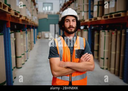 Porträt des Storemans in einem Lager für die Lieferung und den Transport von Industriegütern mit weißem Helm und orange Weste Uniform stehend mit gefaltet Stockfoto