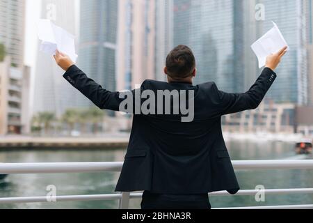Porträt von schönen jungen Geschäftsmann mit erhobenen Händen halten unterzeichnen Vertrag feiern gegen den Blick auf Wolkenkratzer Stockfoto