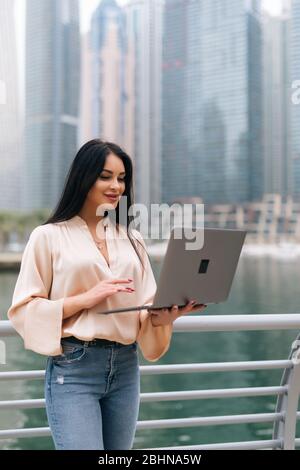 Selbstbewusste Geschäftsfrau, die auf einer Straße in formeller Kleidung steht und einen Laptop hält, während sie mit dem Handy spricht. Stockfoto