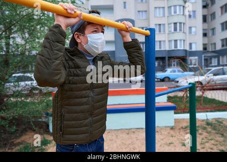 Teenager-Junge steht in der Nähe horizontale Bar von Outdoor-Trainingsgeräten, auf Spielplatz in der Nähe von Hochhäusern mit Wohnungen, eine medizinische Maske auf seinem Gesicht p Stockfoto